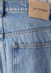 Jasnoniebieskie dżinsy z denimu z skórzaną metką oznaczoną "2Y STUDIOS", widocznym przeszyciem i detalem tylnej kieszeni.