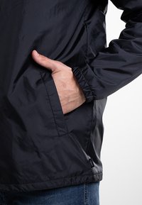 Czarna lekką kurtka z gładką fakturą, elastycznymi mankietami, bocznymi kieszeniami i metką z logo na dolnym końcu, prezentowana na poziomie talii.