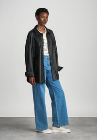 Black leather shirt jacket with two chest pockets, paired with a striped white and cream top and high-waisted blue wide-leg jeans. White slip-on shoes.