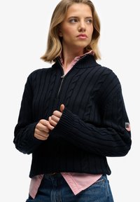 Suéter de punto de cable azul marino con cuello alto, cierre de cremallera en la parte delantera y puños acanalados. Llevado sobre una camisa de color rosa claro con cuello, que presenta un parche de la marca en la manga.