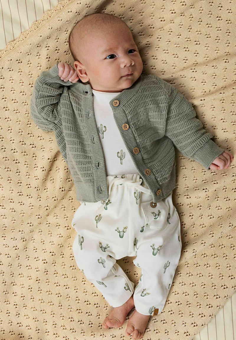 Grüne Strickjacke mit Holzknöpfen, weißes Hemd mit Kaktusmuster und weiche weiße Hose auf einer strukturierten beigen Decke.