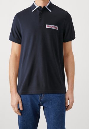 Person trägt ein dunkelblaues Poloshirt mit weiß abgesetztem Kragen und rotem "HILFIGER"-Logo auf der Brust, kombiniert mit blauen Jeans.