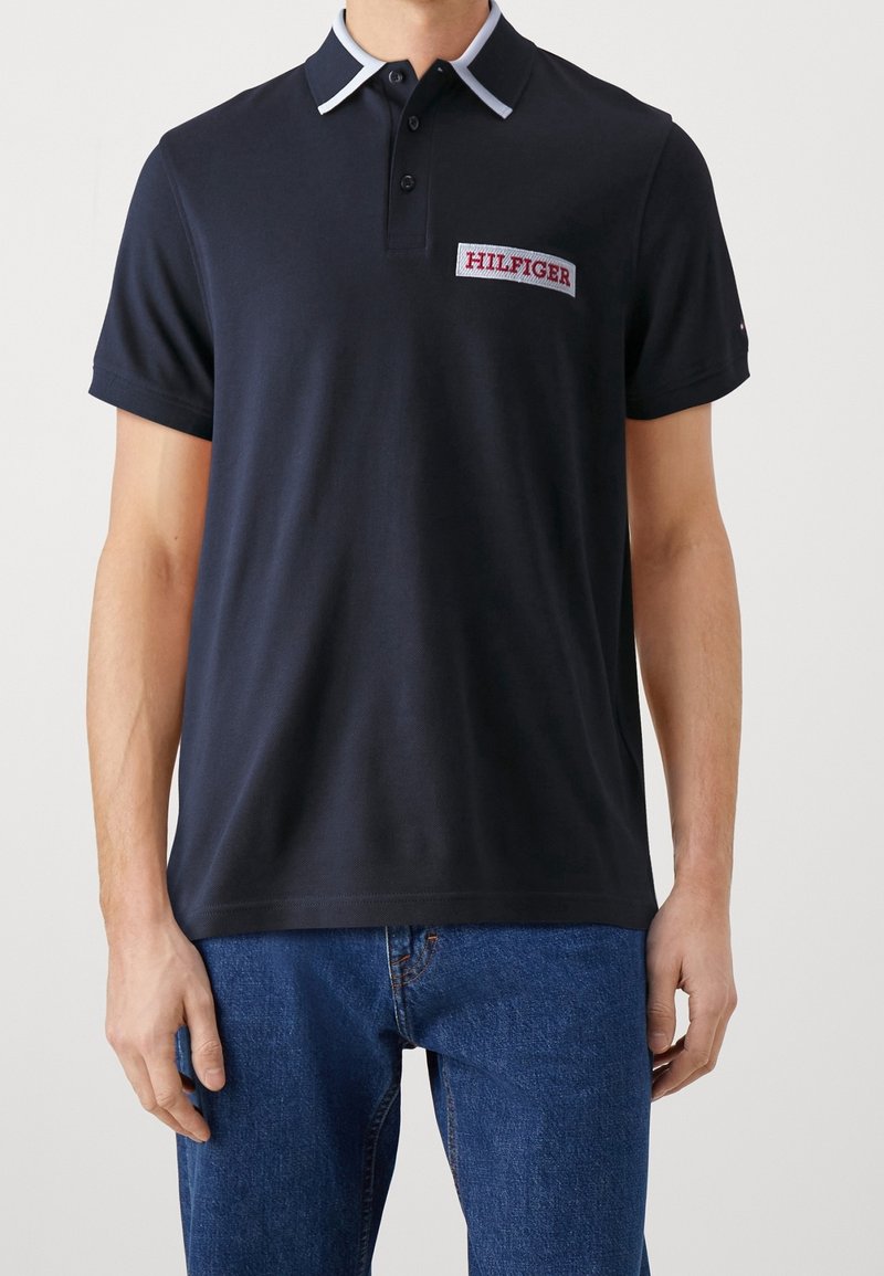 Personne portant un polo bleu marine avec un col bordé de blanc et un logo rouge "HILFIGER" sur la poitrine, associé à un jean bleu.