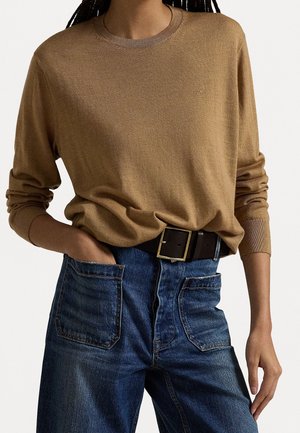 Person iført en beige langærmet sweater stoppet ned i højhåndede blå jeans med store forlommer og et mørkt bælte med spænde i firkantet form.