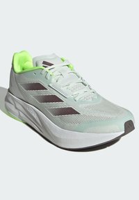 Scarpa da atletica verde chiaro con tomaia in rete testurizzata, accenti marroni scuri, collo imbottito e tecnologia di ammortizzazione visibile sulla suola.
