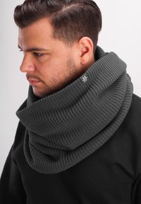 Chunky knit dark gray scarf with a wide, folded design. Features a logo tag on the side. Worn around the neck, showing textured fabric.