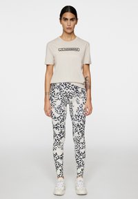 T-shirt in cotone beige con testo nero, abbinato a leggings bianchi con motivi astratti neri. Scarpe da ginnastica bianche completano il look.