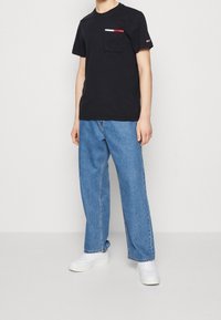 T-shirt en coton noir avec une poche poitrine ornée d'accents rouges et blancs, associé à un jean en denim bleu ample et des baskets blanches.