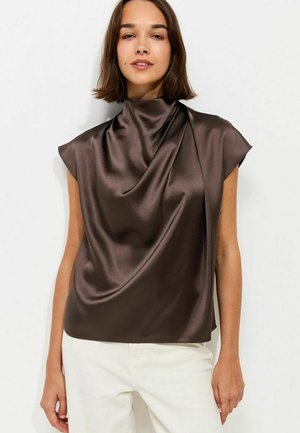 Mujer con una blusa sin mangas de satén marrón oscuro con un escote alto drapeado y pantalones blancos sobre un fondo blanco liso.