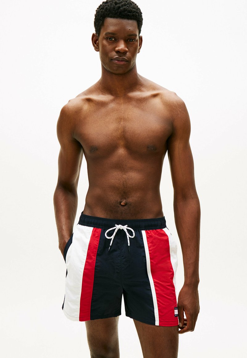 Mężczyzna bez koszulki, stojący z rękami w kieszeniach, ubrany w granatowe, czerwone i białe spodenki kąpielowe z troczkami na jednolitym białym tle.