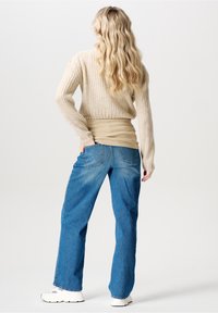 Pull beige en maille avec texture côtelée, associé à un jean bleu clair à jambes droites. Modèle portant des baskets blanches, vu de dos.