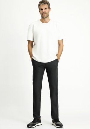 T-shirt bianco a maniche corte abbinato a pantaloni neri slim-fit e scarpe da ginnastica nere, su uno sfondo semplice.