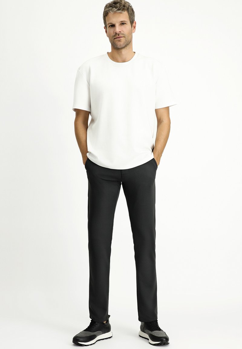 Witte korte mouwen T-shirt gecombineerd met zwarte slim-fit broek en zwarte sportschoenen, tegen een effen achtergrond.