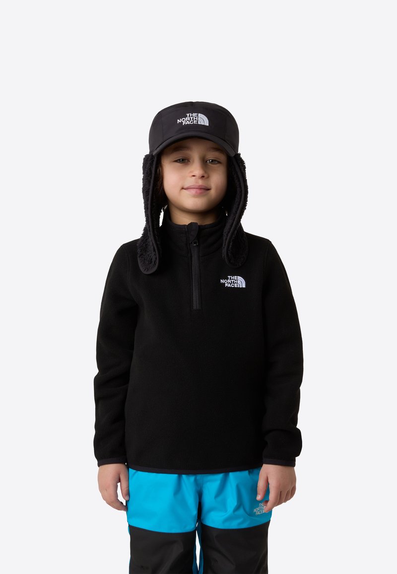 Pulover negru din fleece cu fermoar, asortat cu pantaloni impermeabili albastru aprins și negri. Copilul poartă o șapcă neagră cu urechi acoperite.