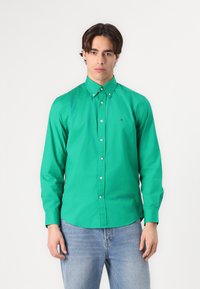 Camicia button-up verde in cotone con colletto a punta, maniche lunghe e un piccolo logo sul lato sinistro del petto. Abbinata a un paio di jeans azzurri chiari.
