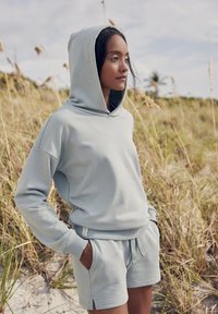 Sudadera azul claro y pantalones cortos a juego en tejido suave. La sudadera presenta una gran capucha, puños acanalados y aberturas laterales; los pantalones cortos incluyen un cordón ajustable.
