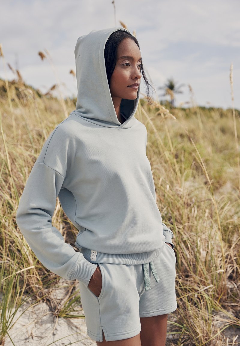 Sudadera azul claro y pantalones cortos a juego en tejido suave. La sudadera presenta una gran capucha, puños acanalados y aberturas laterales; los pantalones cortos incluyen un cordón ajustable.
