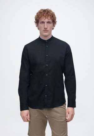 Junger Mann mit lockigem roten Haar, der ein schwarzes Hemd mit Stehkragen und Knöpfen sowie beige Shorts trägt, vor einem schlichten weißen Hintergrund stehend.