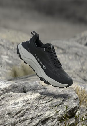 Chaussure de trail running The North Face noire et grise en l'air au-dessus d'un terrain rocheux avec de petites plantes et des particules de terre dispersées.