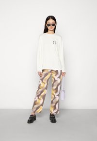T-shirt de manga longa creme com gráfico circular, combinado com calças largas estampadas em tons de castanho, amarelo e branco. Sapatos mocassins pretos e bolsa roxa.