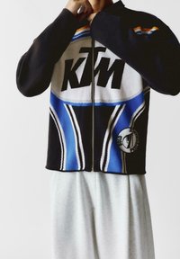 Czarna kurtka z zamkiem błyskawicznym z niebieskimi, białymi i pomarańczowymi wzorami, z dużym logo KTM oraz okrągłą naszywką z przodu na dole.