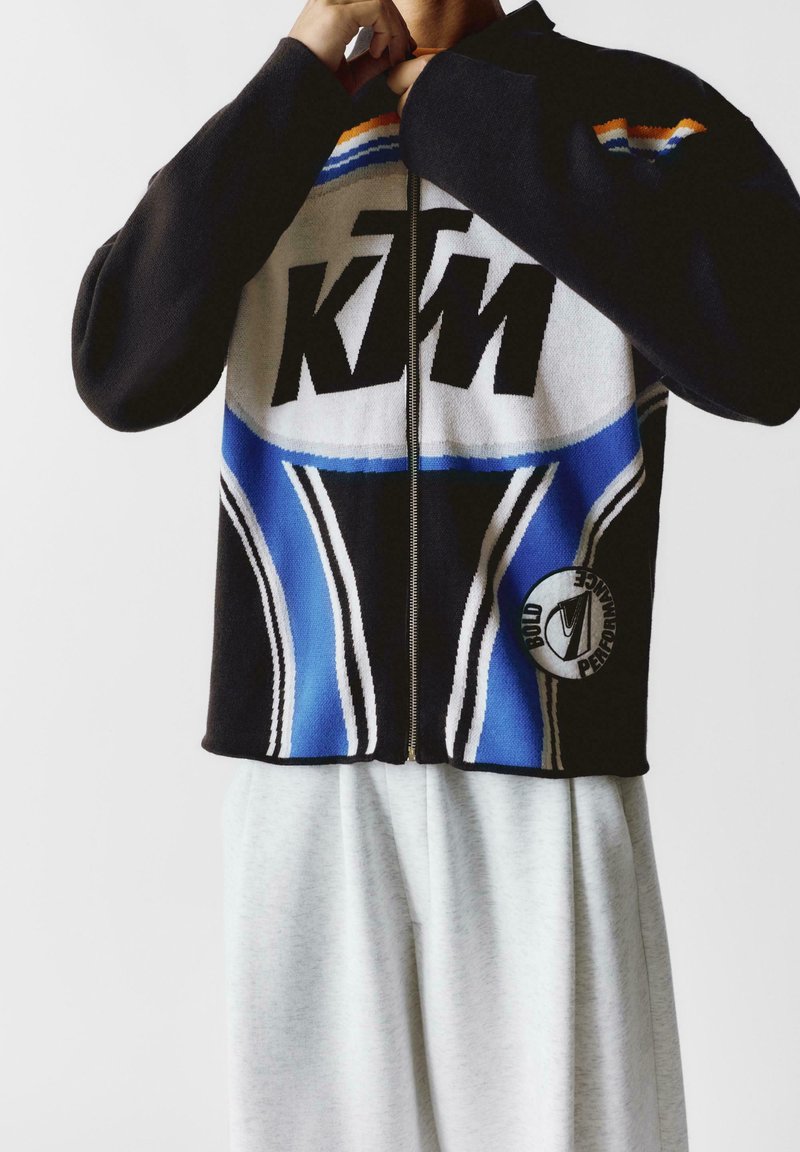 Czarna kurtka z zamkiem błyskawicznym z niebieskimi, białymi i pomarańczowymi wzorami, z dużym logo KTM oraz okrągłą naszywką z przodu na dole.