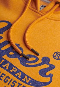 Felpa arancione con un logo blu screpolato e cordini con punte metalliche. Texture morbida e dettagli di design casual.