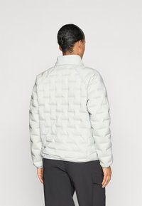 Lys grå quilted puff jakke med høj krave, der har en tekstureret overflade og raglan ærmer, båret over mørke bukser.