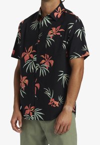 Chemise noire à manches courtes avec un motif floral vibrant en rouge et vert, confectionnée en tissu léger avec une fermeture à boutons sur le devant.