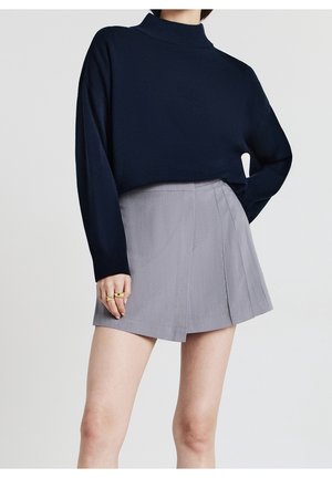 Femme portant un pull à manches longues bleu marine et une mini-jupe plissée gris clair avec des bagues dorées à la main gauche.