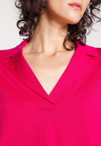 Haut en maille fuchsia avec un col en V et un col pointu. Tissu texturé avec un détail côtelé, mettant en valeur un gros plan sur le col et le col.