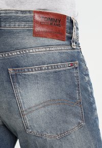 Ljust blå denimjeans med en brun läderetikett där "TOMMY JEANS" är präglat. Inkluderar en bakficka med accentuerad sömnad.