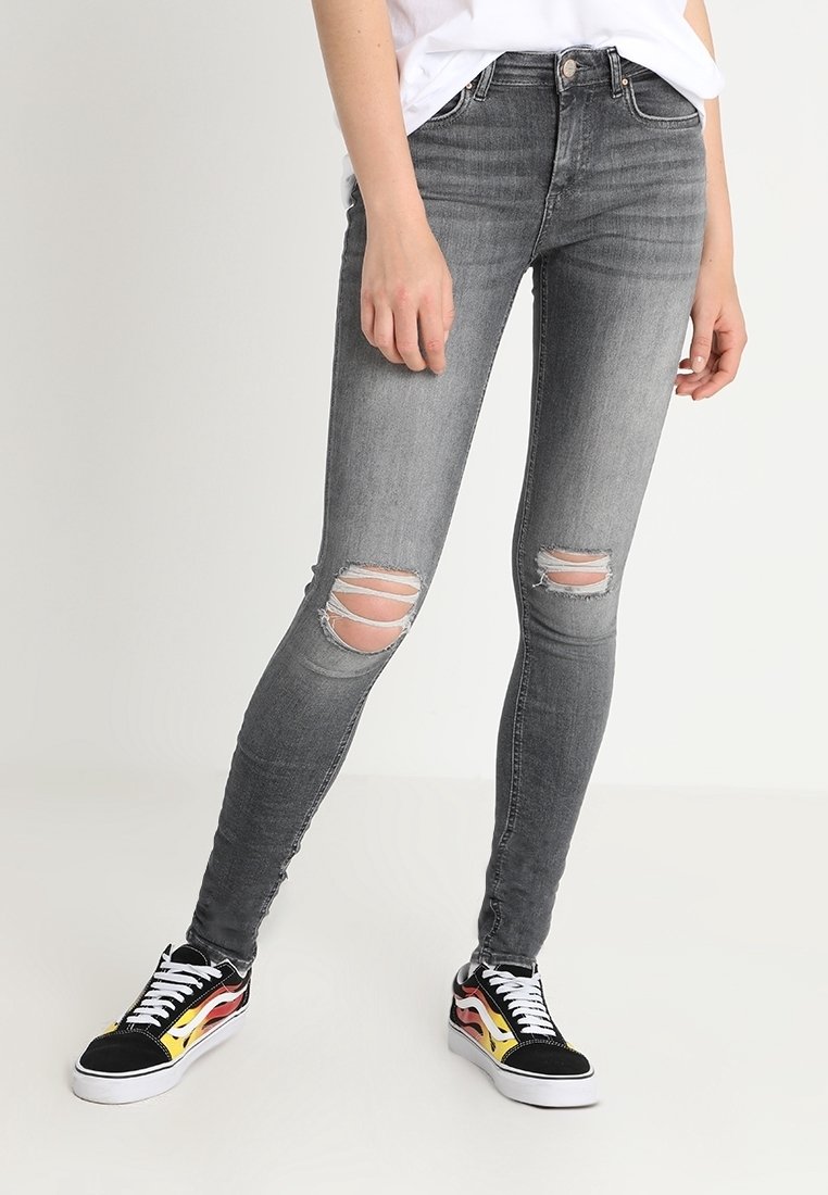 Personne portant un jean skinny gris déchiré et des baskets basses noires avec des motifs de flammes et des lacets blancs, debout sur un fond clair.