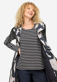 Haut rayé en bleu marine et gris, superposé sous un gilet à motif floral avec fermeture éclair. Présente des cheveux bouclés et des mains dans les poches.