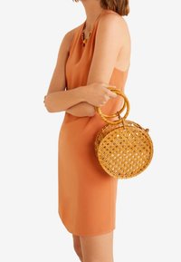 Borsa rotonda intrecciata in rattan naturale di colore marrone chiaro, con due manici circolari. Lo sfondo è di un colore bianco o chiaro uniforme.