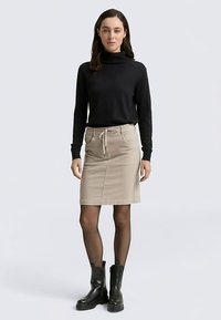 Czarna koszula z długim rękawem i golfem, połączona z beżową spódnicą do kolan z troczkiem w pasie, noszona z czarnymi botkami za kostkę.