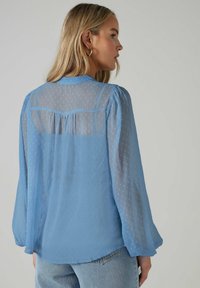 Blusa ligera y azul celeste, semitransparente, con mangas largas abullonadas, que presenta una textura de puntos y un detalle de pliegues en la parte posterior. Combinada con unos vaqueros.
