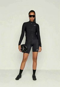 Fekete hosszúujjú kerékpáros mezben és rövidnadrágban lévő nő, fekete sisakot tart a kezében, fekete kerékpáros cipőt, zoknit és nagy napszemüveget visel.