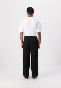 Black trousers feature a straight cut, back pockets, and a small yellow logo on the waistband. Paired with a plain white T-shirt.