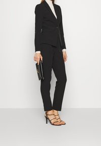 Completo sartoriale nero con giacca a un bottone e pantaloni slim-fit. Indossato con un dolcevita color crema e tacchi neri a cinturino, mentre si tiene una pochette.