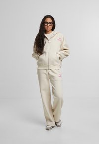 Beige zipe-uppaná mikina a zodpovedajúce teplákové nohavice z mäkkej látky. Mikina má na hrudi ružové logo. Model bol postavený na neutrálnej pozadí.