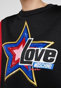 Mustapaita, jossa on värikäs tähtigrafiikka sinisessä, punaisessa ja vaaleanpunaisessa, jossa on paljetteja, sekä valkoinen logo-teksti "Love Moschino Team."