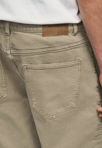 Beige denim pants with a textured finish, featuring two back pockets, visible stitching, and a leather label that reads "DENIM SUPPLY Co."