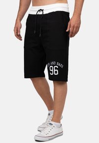 Shorts noirs avec une ceinture élastique blanche et un cordon de serrage, comportant des poches latérales. Le design inclut le numéro imprimé "96" et le nom de la marque en blanc.