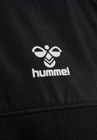 Tessuto nero con un motivo testurizzato. Presenta un logo bianco di un'ape stilizzata e il testo "hummel", bordi cuciti e una texture morbida.