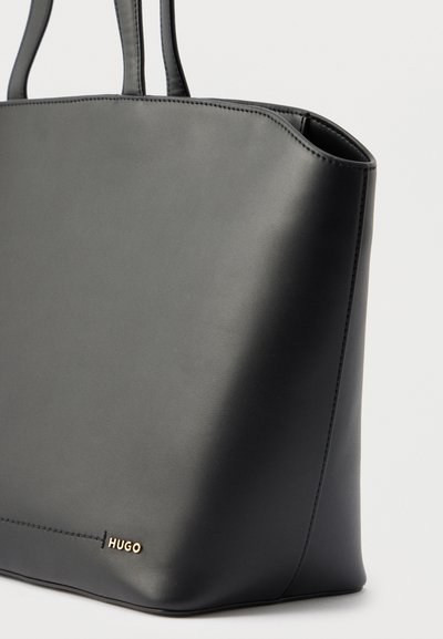 Sac cabas en cuir noir avec deux poignées et petit logo doré "HUGO" dans le coin inférieur avant, design minimaliste.