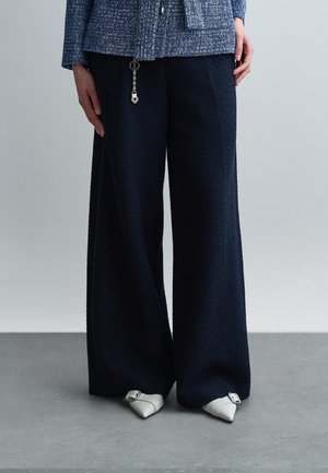 Osoba ubrana w granatowe spodnie o szerokich nogawkach, niebieską teksturowaną kurtkę z detalem w postaci breloczka oraz białe szpiczaste buty z srebrnymi klamrami.