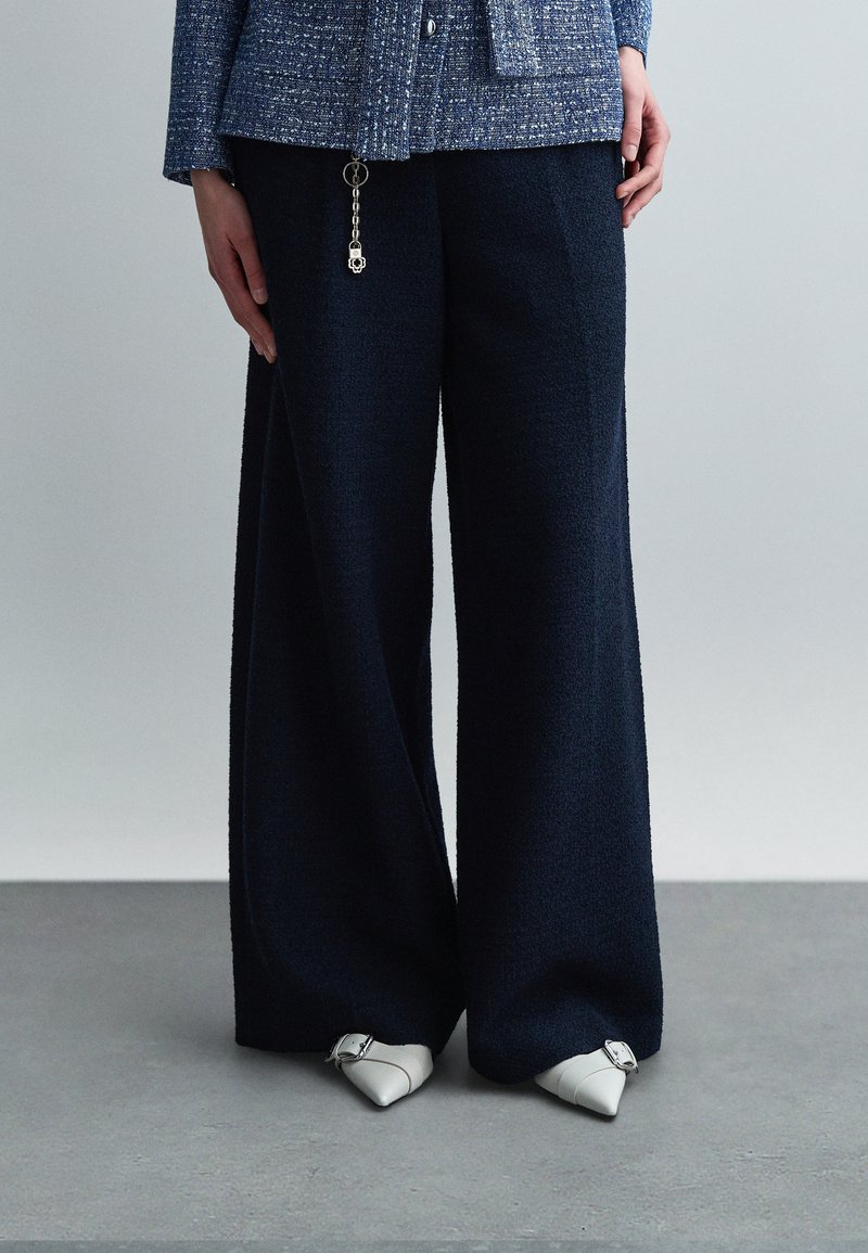 Άτομο που φοράει ναυτικό μπλε παντελόνι με φαρδιά μπατζάκια, μπλε υφασμάτινο σακάκι με λεπτομέρεια μπρελόκ, και λευκά μυτερά παπούτσια με ασημένιες αγκράφες.