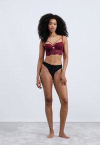 Burgundy lace bralette dengan tali yang dapat disesuaikan dan potongan sentral, dipadukan dengan underwear hitam potongan tinggi di latar belakang yang terang.