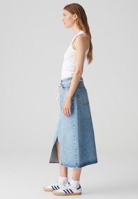 Gonna maxi in denim blu chiaro, con spacco frontale, vita aderente e chiusura con cordoncino. Abbinata a una canotta bianca e sneakers.